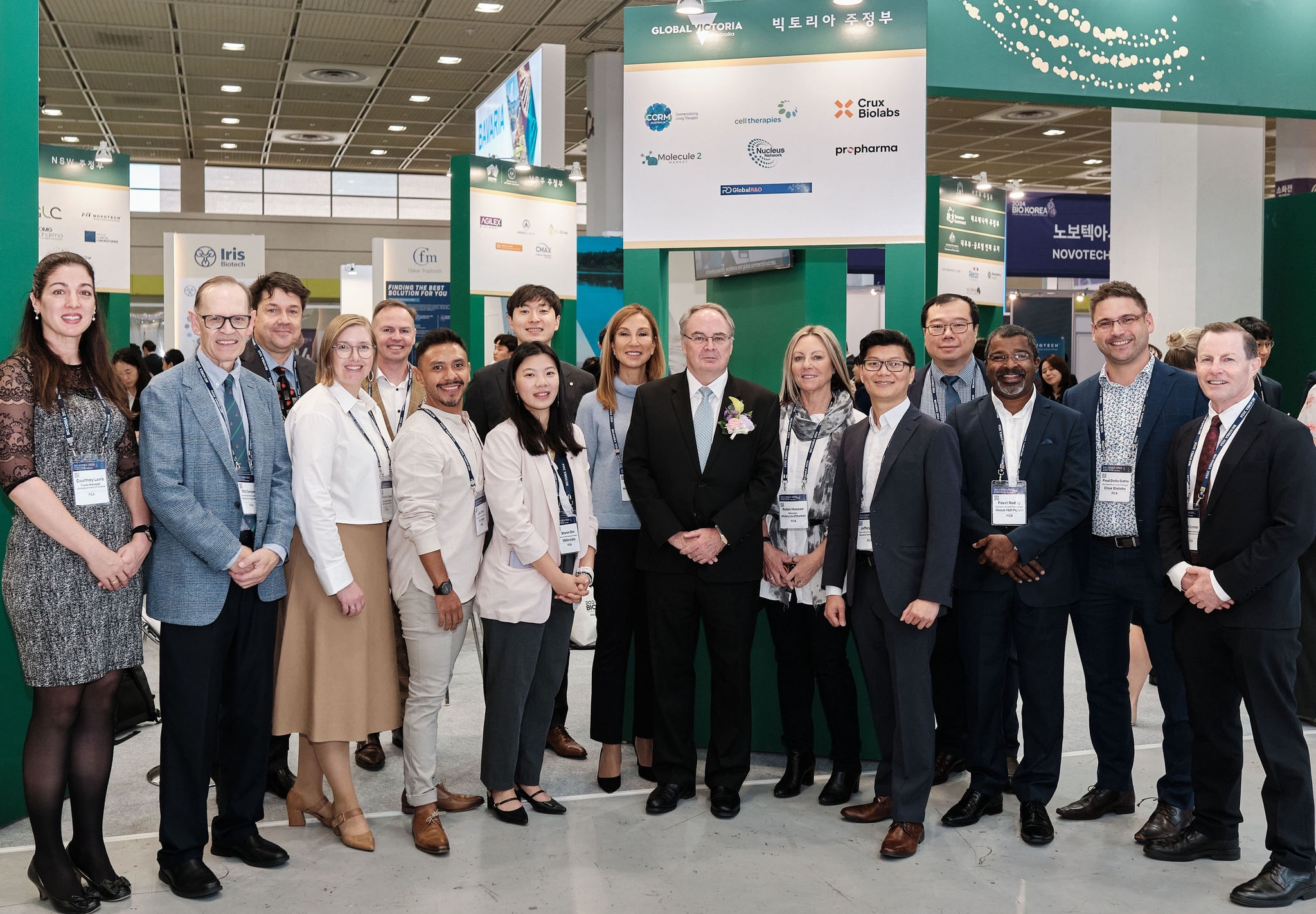Sixteen people dressed in business attire at a tradeshow, smiling in front of signs with company logos.