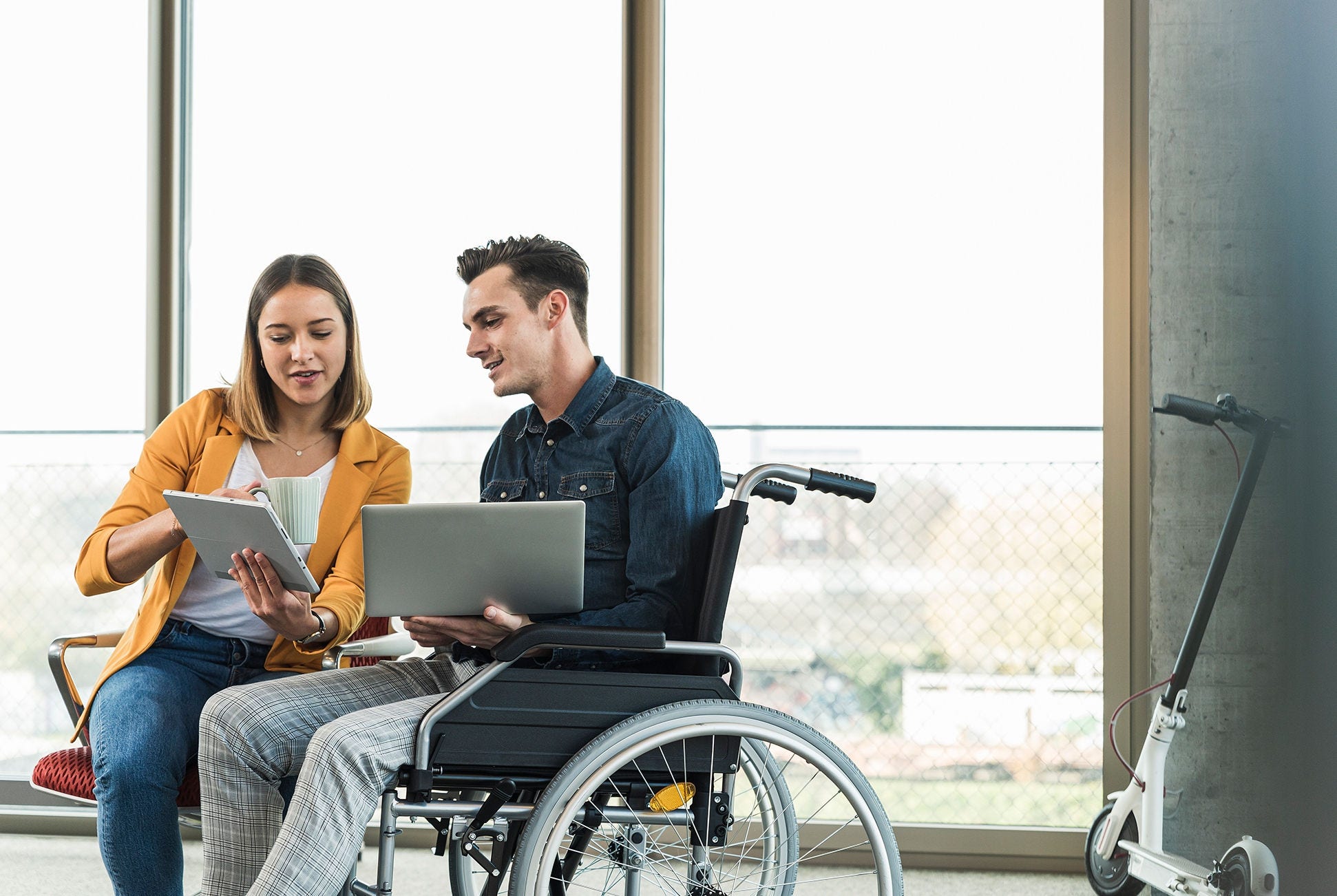 Young businessman with laptop in wheelchair and businesswoman with tablet in office