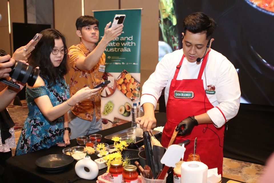 A cooking demonstration by a chef in a red apron while onlookers take video and photos