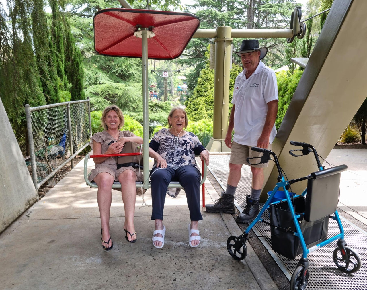 Two women sitting in a chairlift. A man stands on the right. A mobility aid is in the foreground.