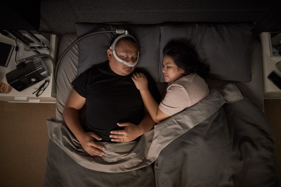 A man and a woman sleeping in bed. The man is wearing ResMed's sleep apnoea device.