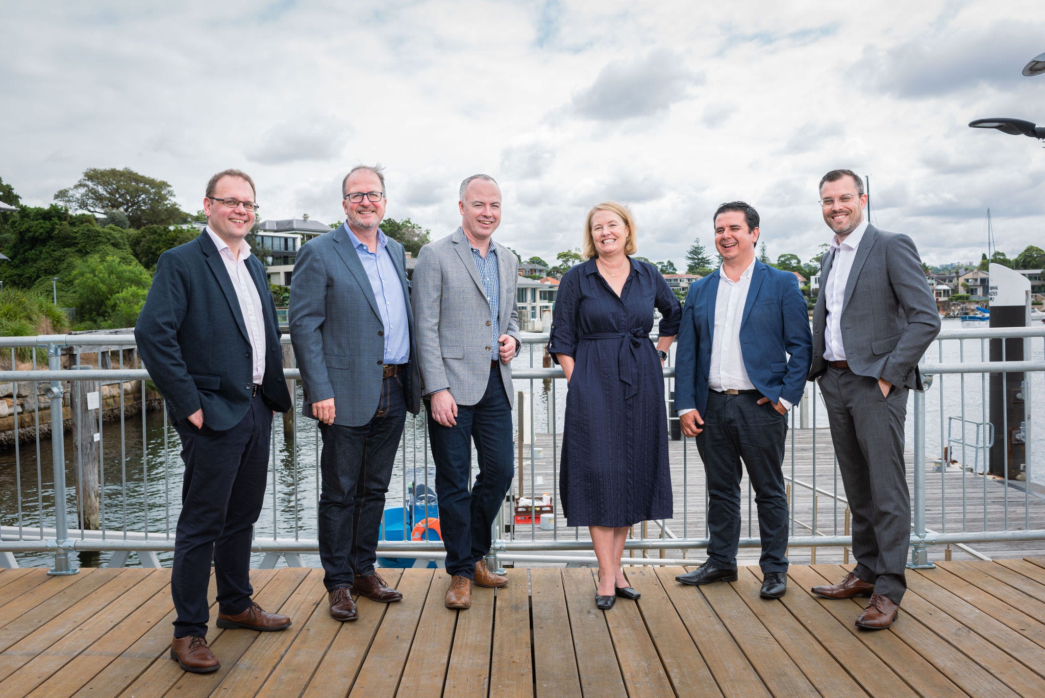 Six professionals dressed in corporate attire, smiling on a wharf on a cloudy day.
